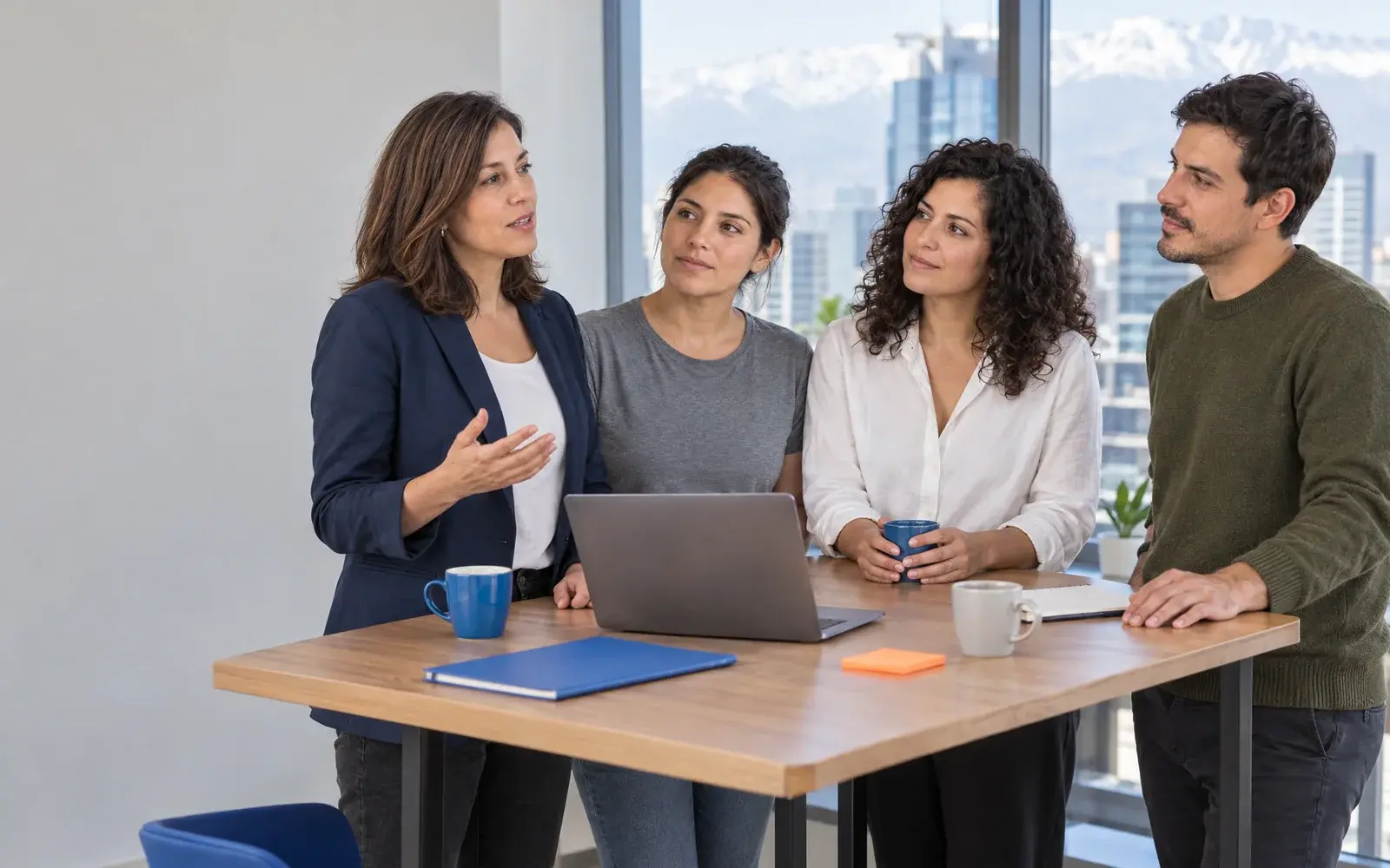 Cuatro personas conversan de pie alrededor de una mesa con una laptop.